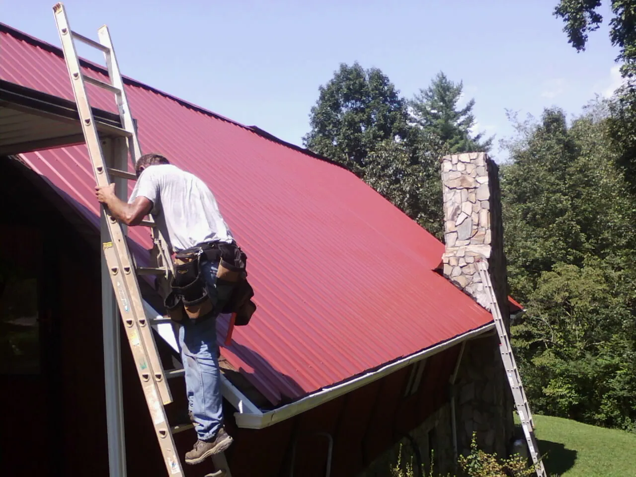 Roofer on a ladder beside a red metal roof and stone chimney — Soffit & Fascia Repair by Roofing Brothers in Marengo